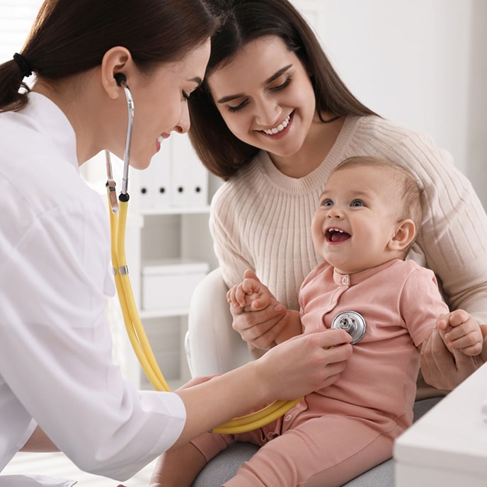 A pediatrician using a stethoscope to examine a smiling baby sitting on her mother's lap.