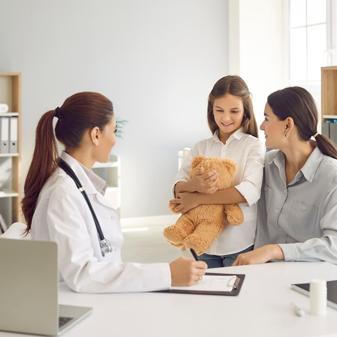 A young female doctor in her office talking to a woman and her daughter who holds a teddy bear in her arms.