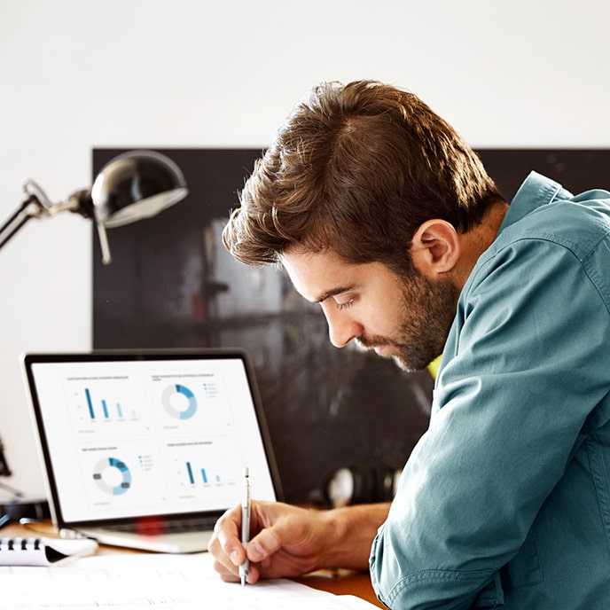 Mężczyzna siedzący przy stole porównuje dane z raportu z wykresami na swoim komputerze przenośnym.
